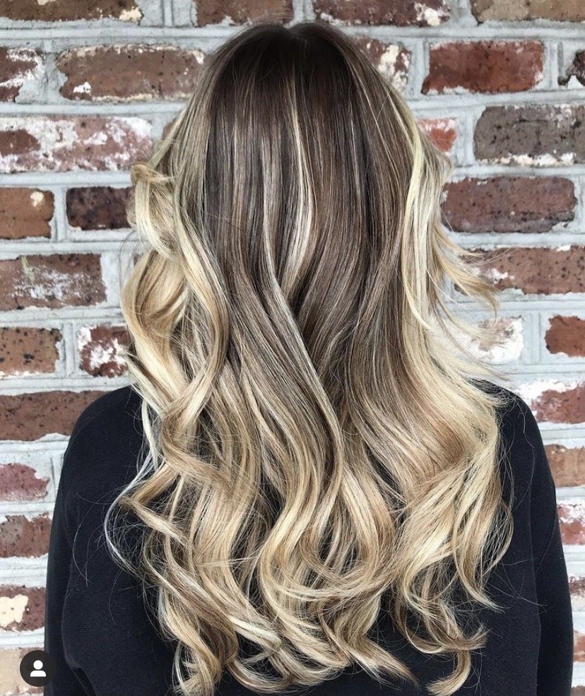 Blonde balayage in loose curls, photographed against the salon’s brick wall